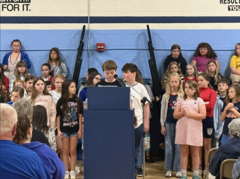A student stands at a podium reading during the Memorial Day program while classmates stand behind them and audience members watch.