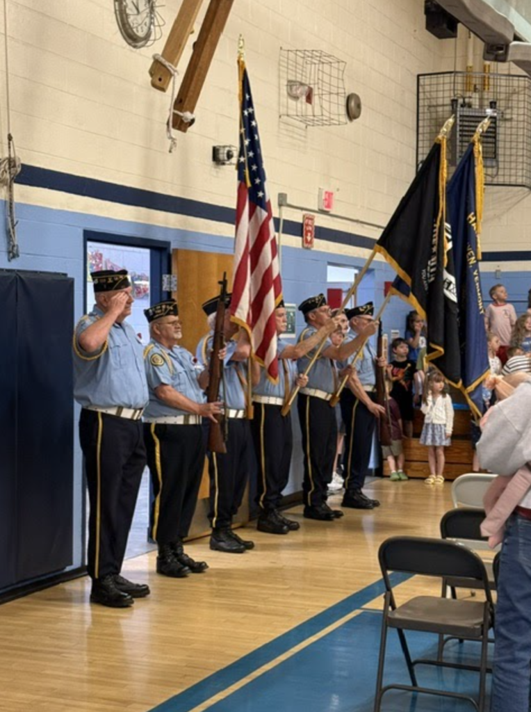 Members of the local Color Guard salute and present flags during the Memorial Day ceremony as students and families stand and observe.