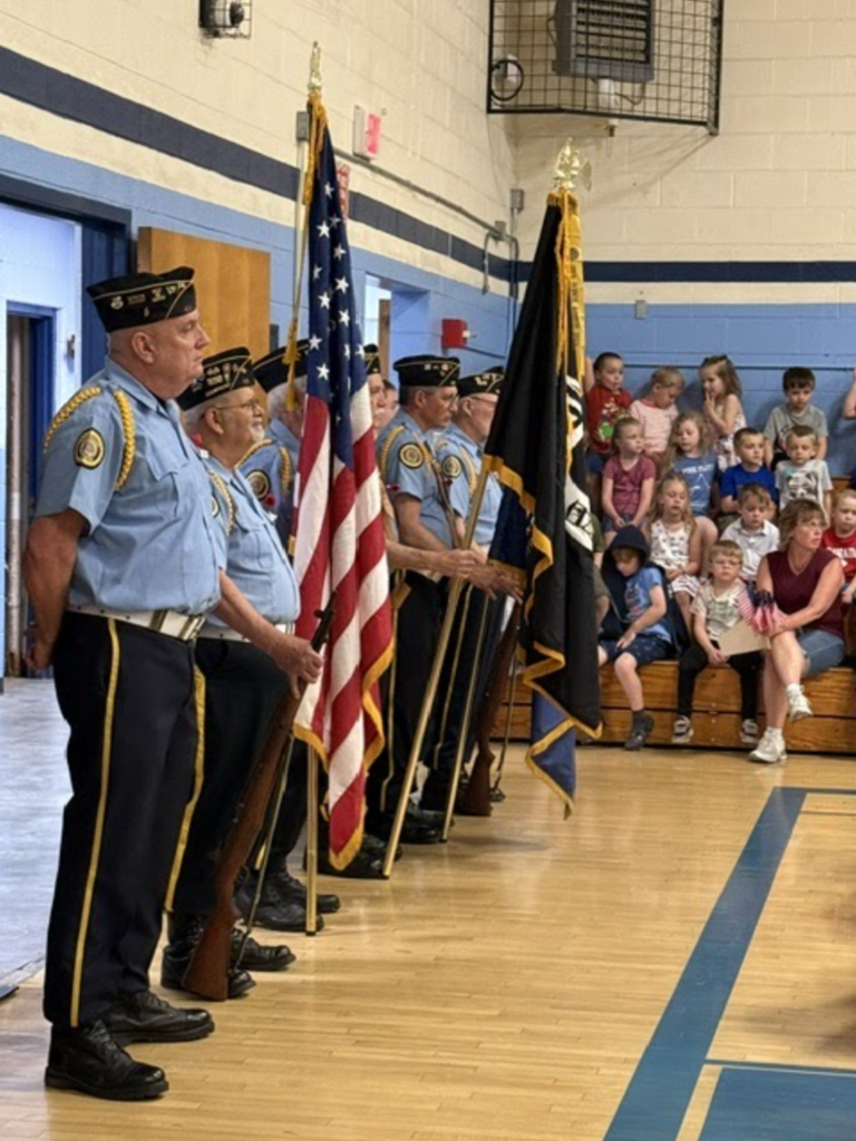 Members of a local Color Guard unit stand in formation inside the Fair Haven Grade School gymnasium holding flags and ceremonial rifles while students and families watch during a Memorial Day program.