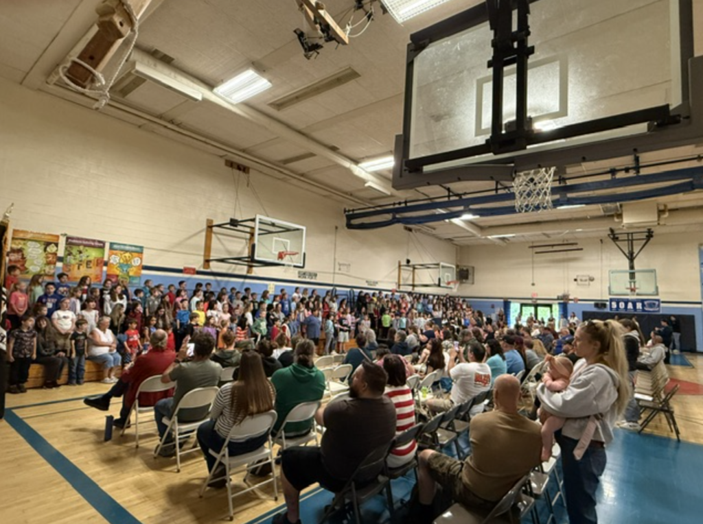 A wide view of the Fair Haven Grade School gym shows students performing during the Memorial Day program while families and community members sit and watch.