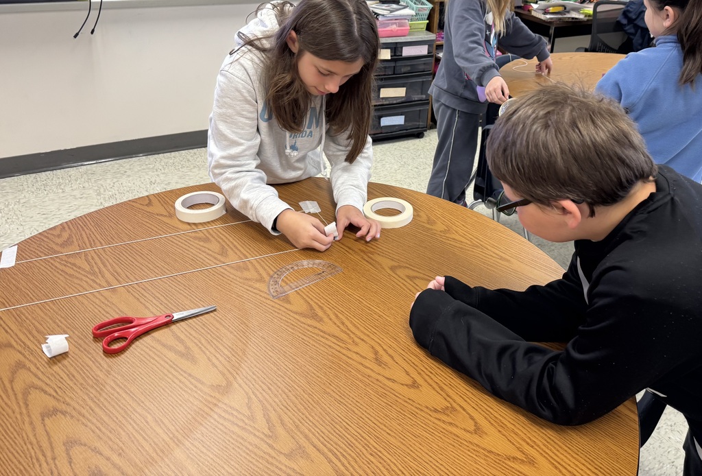 A close-up shot of a classroom table where a young girl in a gray hoodie is measuring and taping two parallel lengths of white string onto the surface. A boy wearing glasses and a black sweatshirt leans forward, watching her progress attentively. A red pair of scissors, rolls of masking tape, and a clear plastic protractor rest on the wooden tabletop between them.