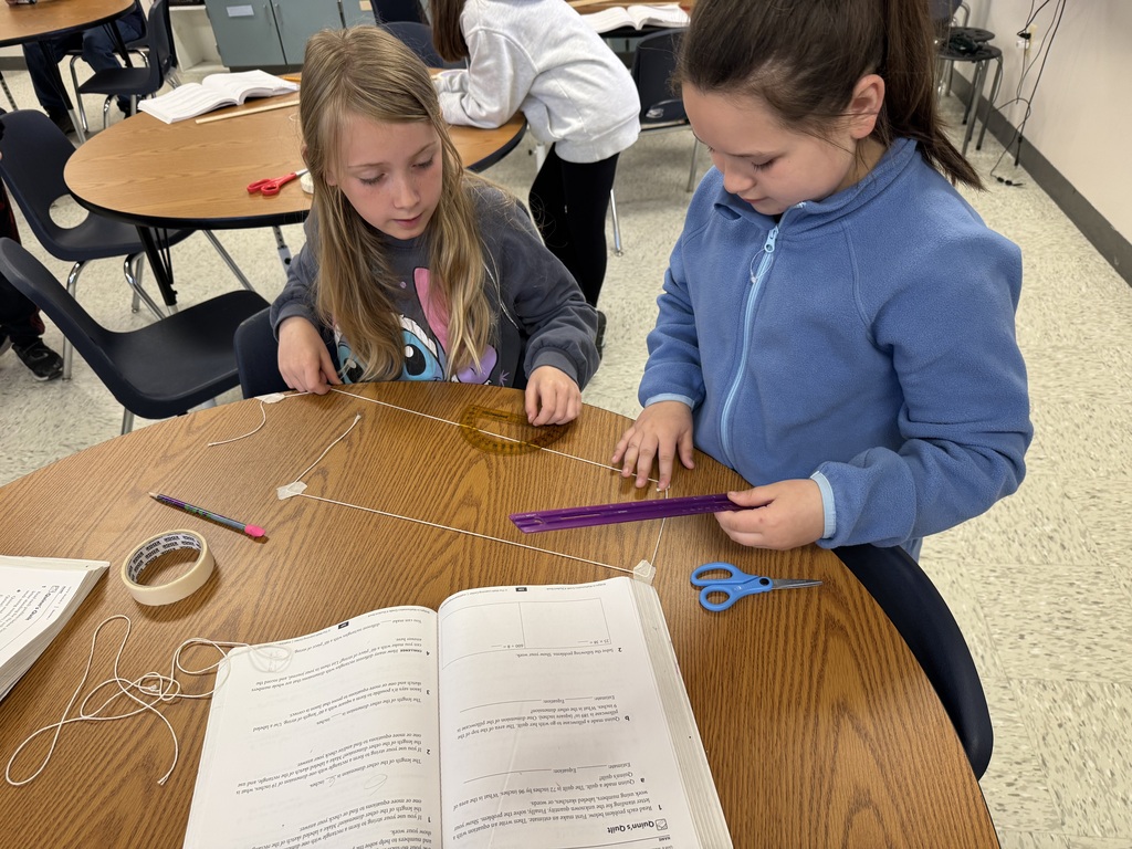 Two young girls collaborate at a round wooden table, working on a geometry problem. They have used white string and masking tape to construct a perfect rectangle on the table's surface. One girl uses a clear protractor to check the angles, while the other holds a purple ruler. An open math workbook sits in the foreground, showing equations and word problems related to perimeter and area.