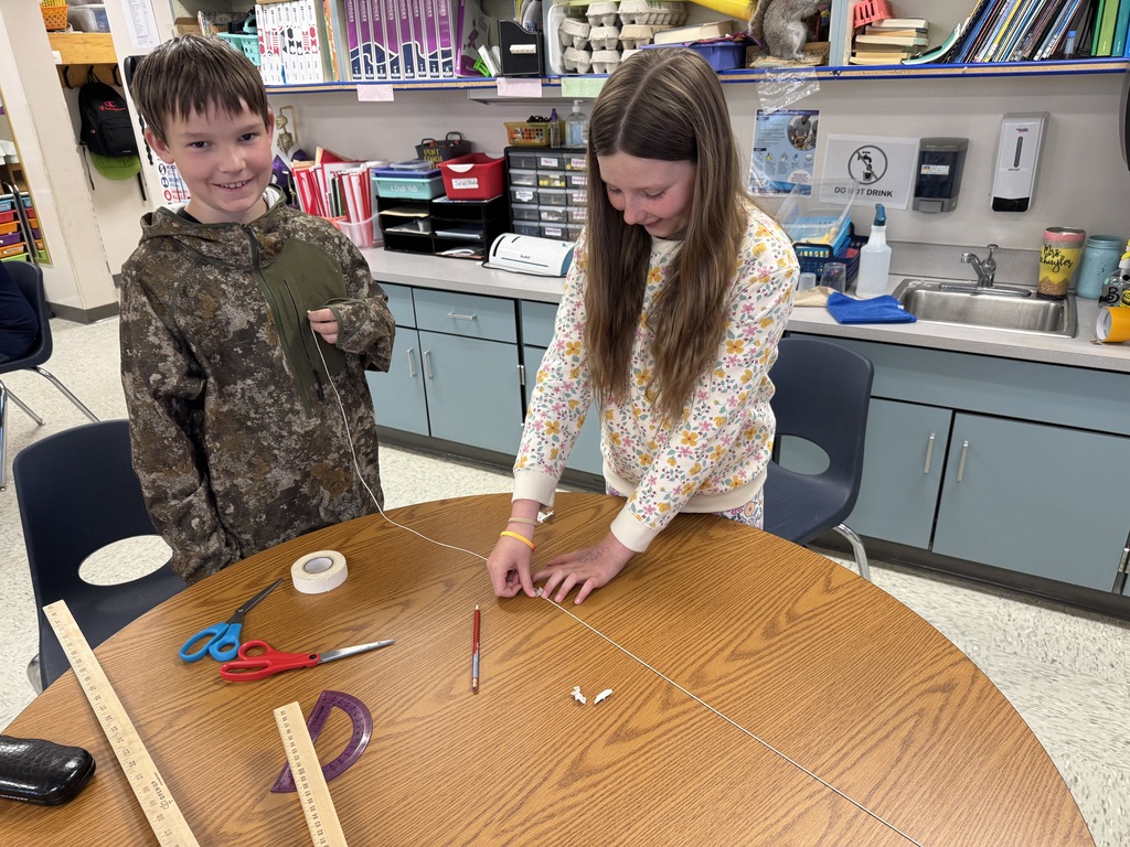 A boy and a girl work together at a round wooden classroom table during a hands-on math lesson. The boy, wearing a camouflage hoodie, smiles at the camera while holding one end of a long piece of white string. The girl, in a floral sweatshirt, carefully tapes the string down onto the tabletop. Scattered across the table are rulers, a protractor, scissors, masking tape, and a pencil, with classroom shelving and a sink visible in the background.