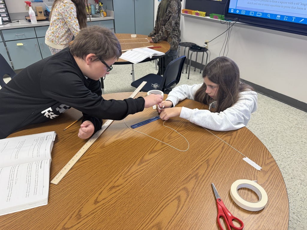 A close-up shot of a classroom table where a young girl in a gray hoodie is measuring and taping two parallel lengths of white string onto the surface. A boy wearing glasses and a black sweatshirt leans forward, watching her progress attentively. A red pair of scissors, rolls of masking tape, and a clear plastic protractor rest on the wooden tabletop between them.