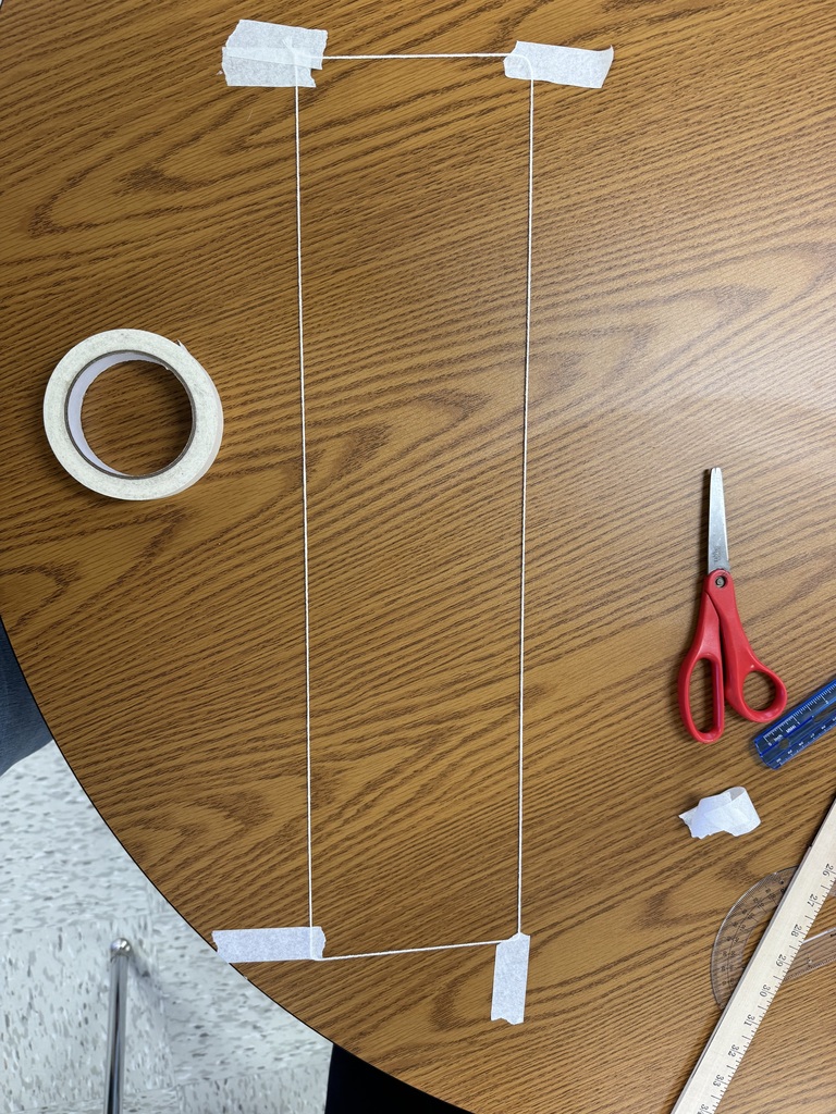 A top-down, bird's-eye view of a geometric shape constructed on a wooden classroom table. A long piece of white string has been arranged and secured at four corners with pieces of white masking tape to form a narrow rectangle. Surrounding the shape on the table are a roll of masking tape, a pair of red scissors, a blue ruler, a protractor, and a wooden yardstick.