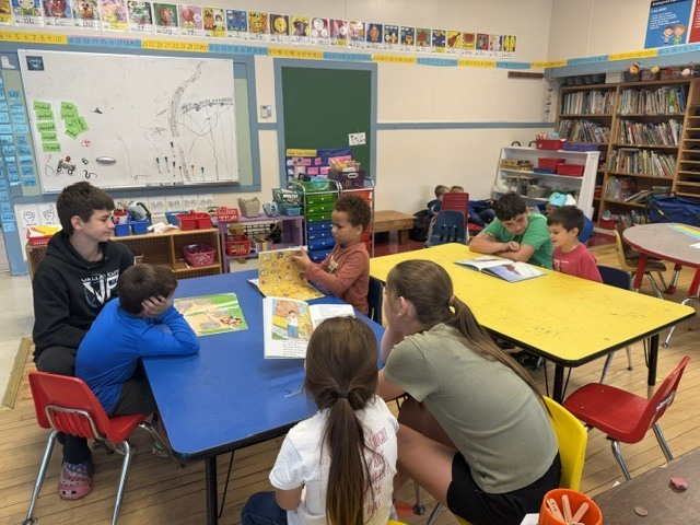 Students sit around colorful classroom tables reading together in small groups while older student readers share books with younger students during a classroom literacy activity.
