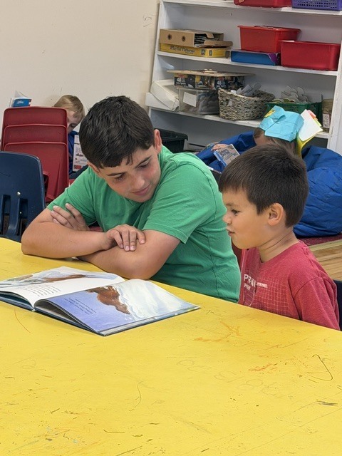 An older student and a younger student sit together at a yellow classroom table reading a picture book and discussing the story.