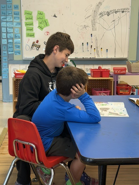 An older student and a younger student sit side by side at a blue classroom table reading a book together while classroom materials and a whiteboard are visible in the background.