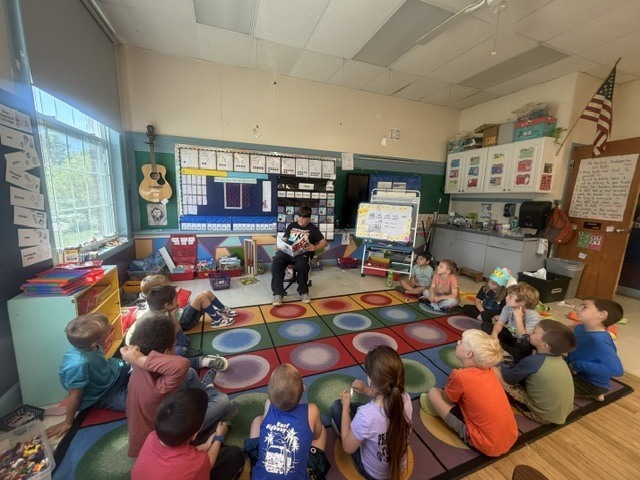 Students sit in a circle on a colorful classroom rug listening as an older student reads aloud at the front of the room during a classroom reading activity.