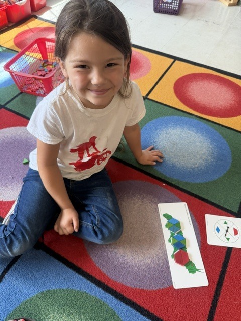 A smiling student sits on a colorful classroom rug next to a completed pattern-building activity made with geometric shapes.