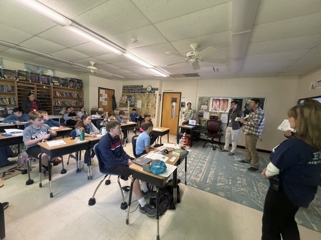A classroom presentation is underway as students listen attentively while multiple adults stand at the front speaking and holding materials for discussion.