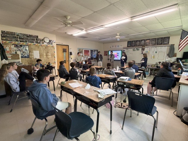Students sit at individual desks in a classroom listening to several adults presenting at the front of the room near a display screen during a classroom presentation.