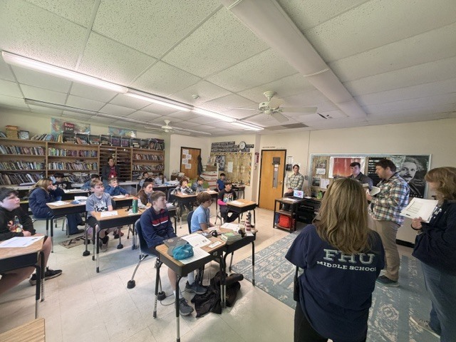Students seated at desks face the front of a classroom as presenters speak and share information. Bookshelves, posters, and classroom materials line the walls.