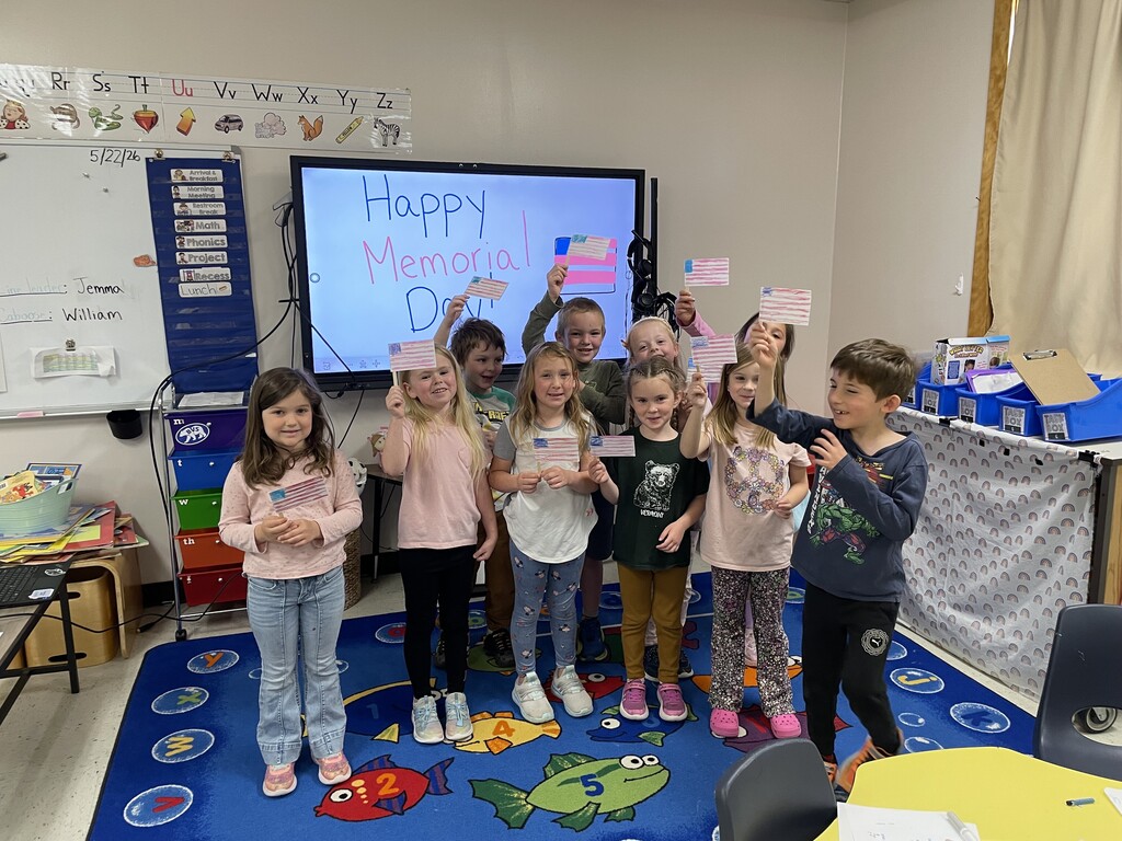 An image of a classroom of young students holding up handmade American flags. In the center, a large digital screen reads "Happy Memorial Day!". On the left is a whiteboard with a list of daily tasks and the alphabet overhead. The room is decorated with a colorful rug, a table, and bins.
