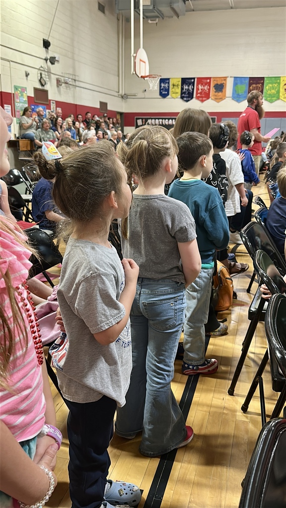 Students stand with hands over their hearts during the Memorial Day assembly while teachers and families observe in the background.