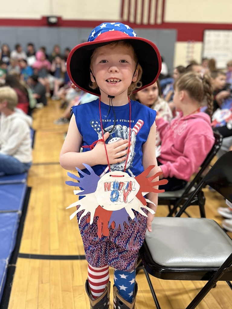 A student dressed in patriotic red, white, and blue clothing and a star-spangled hat stands smiling during Castleton Elementary School’s Memorial Day assembly while placing a hand over their heart.