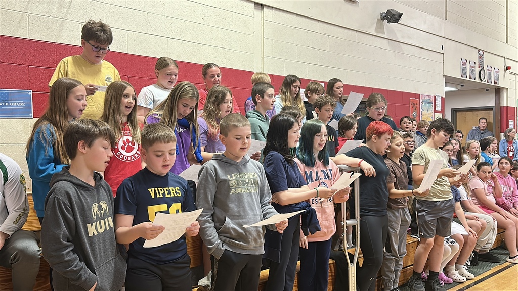 Students stand on risers and bleachers holding papers while singing together during the school’s Memorial Day program.