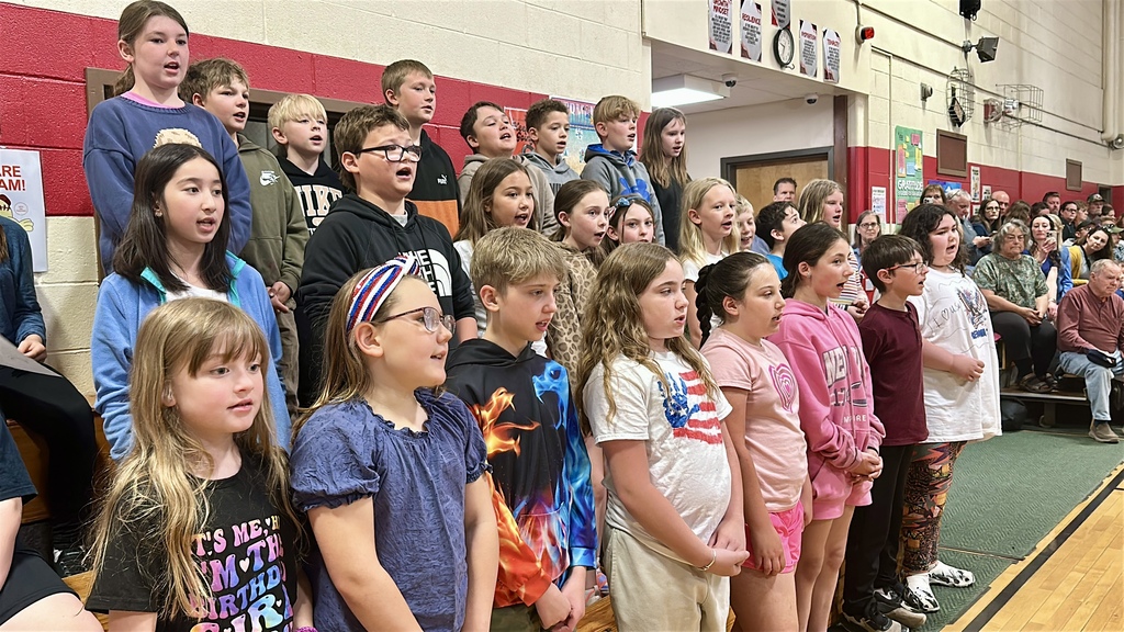 Students stand together singing during the Memorial Day assembly as families and community members watch from the audience.