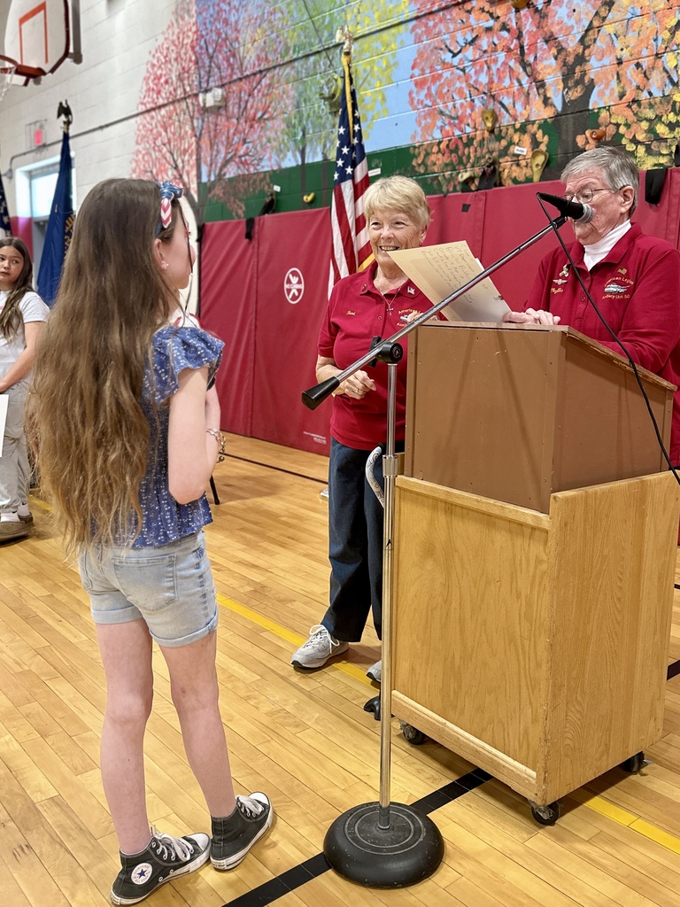 A student stands at a podium receiving recognition during the American Legion Auxiliary contest award presentation while two Auxiliary members smile nearby.