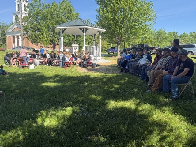 Community members and veterans sit in folding chairs under the shade of trees while listening to a student band perform near a gazebo.