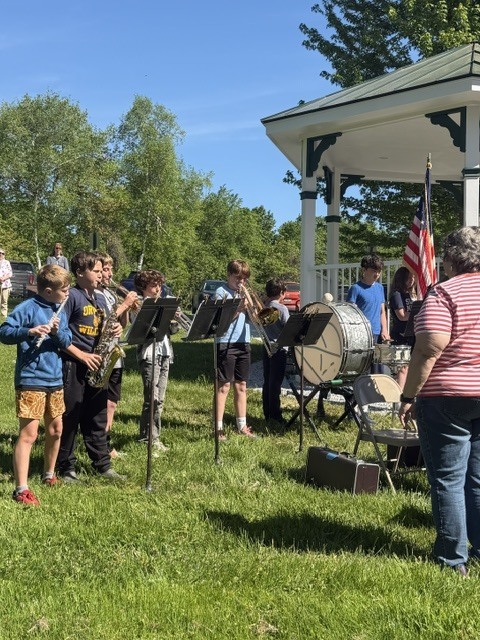 Student musicians stand and play instruments outdoors near a gazebo during a Memorial Day ceremony.