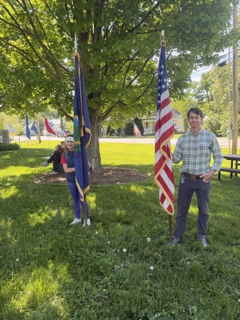 A student and a teacher stand holding the American flag and a state flag beneath a large tree during a Memorial Day ceremony.