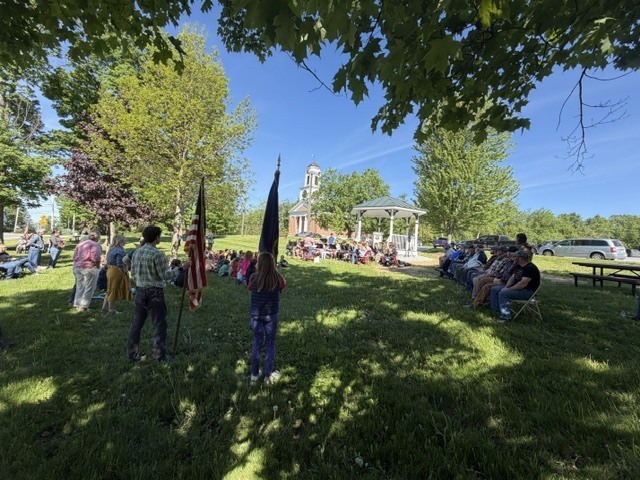 A wide view of an outdoor Memorial Day ceremony shows students holding flags, community members seated under trees, and a gazebo at the center of the event.