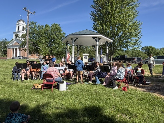 Students from a school band sit outdoors near a gazebo performing music during a Memorial Day gathering, with a church visible in the background on a sunny day.