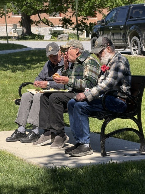 Three veterans sit together on a park bench enjoying food and conversation during a community Memorial Day gathering.