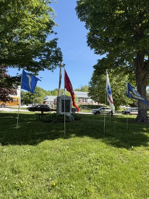 Military service flags stand around a memorial stone beneath large trees on a bright sunny day.