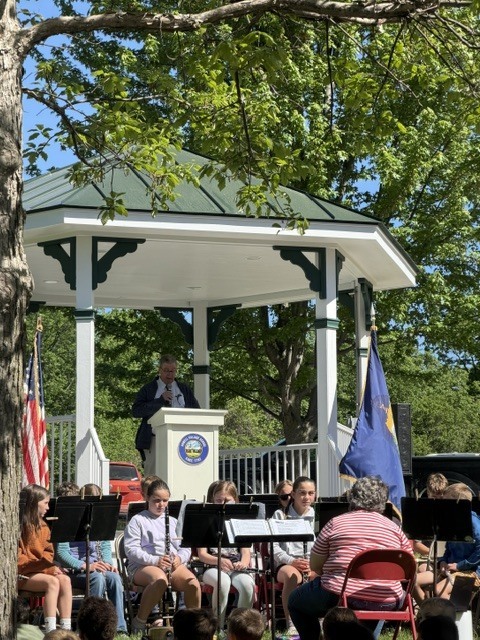 A speaker stands at a podium inside a gazebo addressing attendees while student musicians sit nearby during an outdoor Memorial Day event.