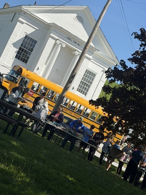 People gather around tables serving food near a town hall building and a school bus during a community Memorial Day event.