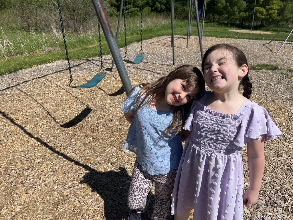  Two young girls are posing playfully together on a sunny playground covered in woodchips. The girl on the left tilts her head smilingly, wearing a light blue lace top and leopard-print pants. The girl on the right makes a goofy, wide-toothed grimace at the camera, sporting a braided ponytail and a light purple ruffled dress with pom-pom textures. Empty swings are visible in the background.  
