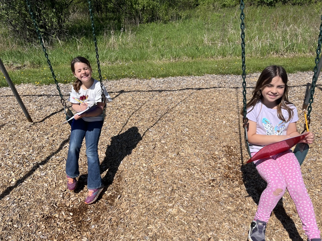  Two young girls sit side-by-side on green playground swings over woodchips, smiling at the camera. The girl on the left wears a white t-shirt, blue jeans, and purple sneakers, and holds a clipboard and pencil. The girl on the right has long brown hair, wears a light purple shirt with a butterfly graphic, pink patterned leggings, and is holding a red folder and a pencil. A grassy treeline is behind them. 