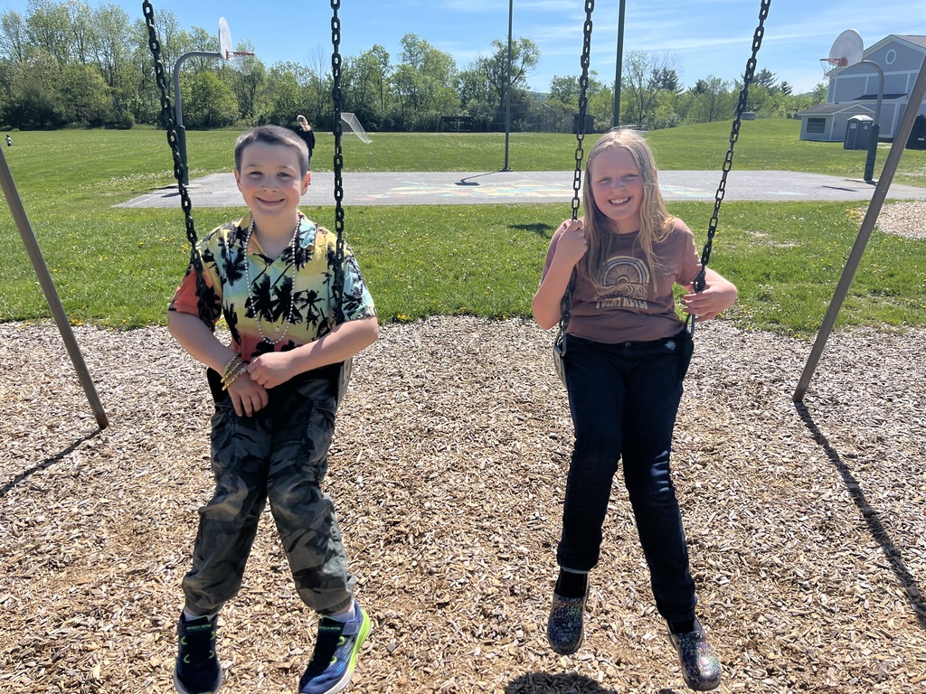  A boy and a girl smile while sitting next to each other on playground swings on a bright day. The boy on the left wears a colorful short-sleeve button-down shirt with a palm tree pattern, camouflage pants, a beaded necklace, and black sneakers. The girl on the right has long blonde hair and wears a brown graphic t-shirt, dark jeans, and multi-colored shoes. An open green field with basketball hoops is behind them. 