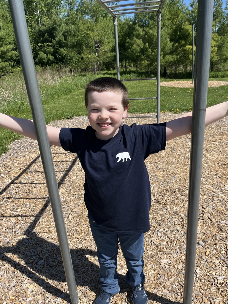 A young boy with a short buzz cut stands smiling on a woodchip-covered playground on a sunny day. He is wearing a dark blue t-shirt with a small white bear logo on the left chest, blue jeans, and blue sneakers. His arms are outstretched horizontally, lightly touching two vertical metal poles of a monkey bar set structure. Green trees and a grassy hill are visible in the background.  