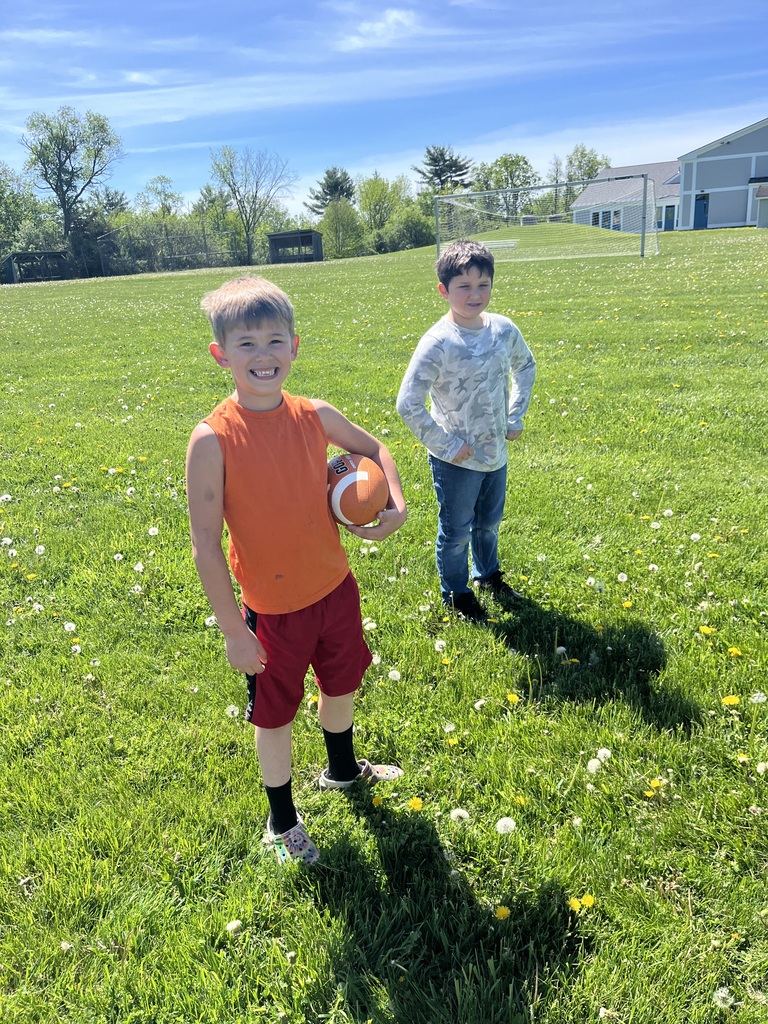 Two boys stand in a large, vibrant green field filled with yellow dandelions under a bright blue sky. The boy in the foreground smiles broadly at the camera while holding an orange football under his arm, wearing an orange sleeveless shirt, red athletic shorts, black socks, and patterned clogs. The boy in the background stands with his hands on his hips, wearing a light-colored camouflage long-sleeve shirt and blue jeans. A soccer goal stands further back in the fiel