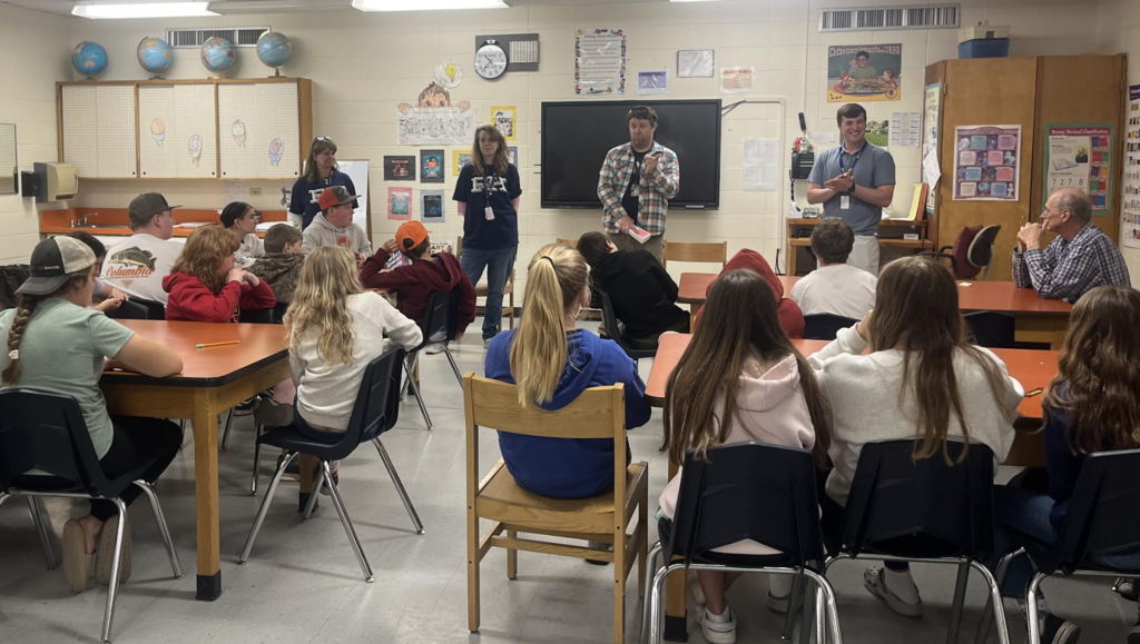 Middle school administrators and faculty members stand at the front of a classroom speaking with a group of sixth grade students seated around tables. Students listen attentively during a visit focused on preparing them for the transition to middle school. A smart board, classroom posters, and learning materials are visible in the background.