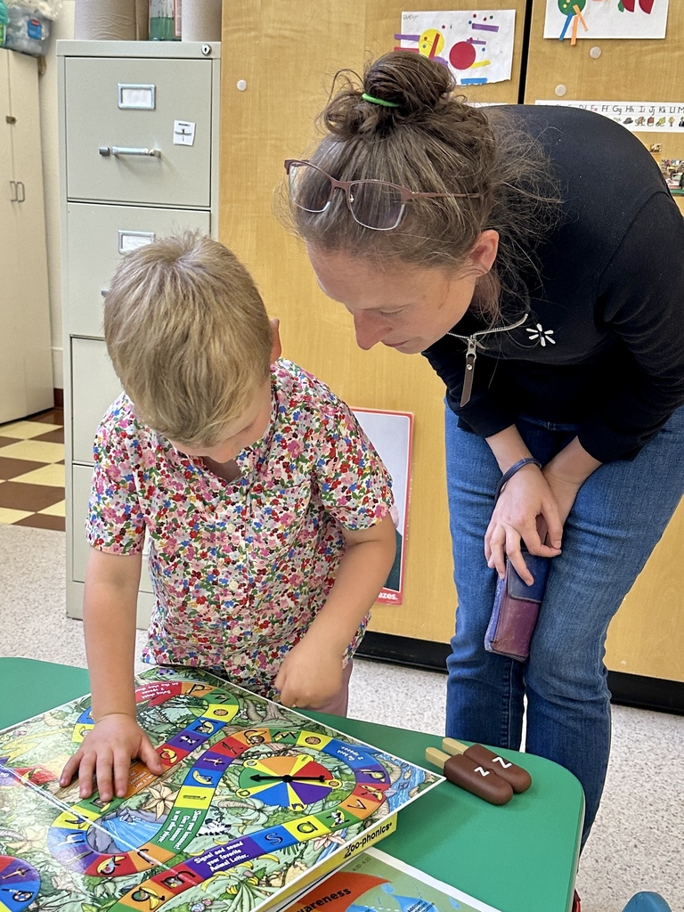An adult leans down beside a student as they look at and discuss an educational game on a classroom table.