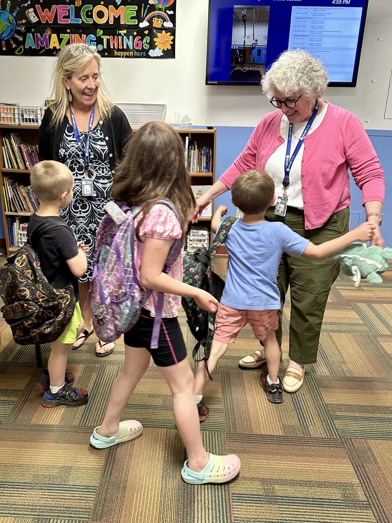 Two school staff members smile and greet students wearing backpacks as they arrive for Celebration of Learning in a school hallway and library area.