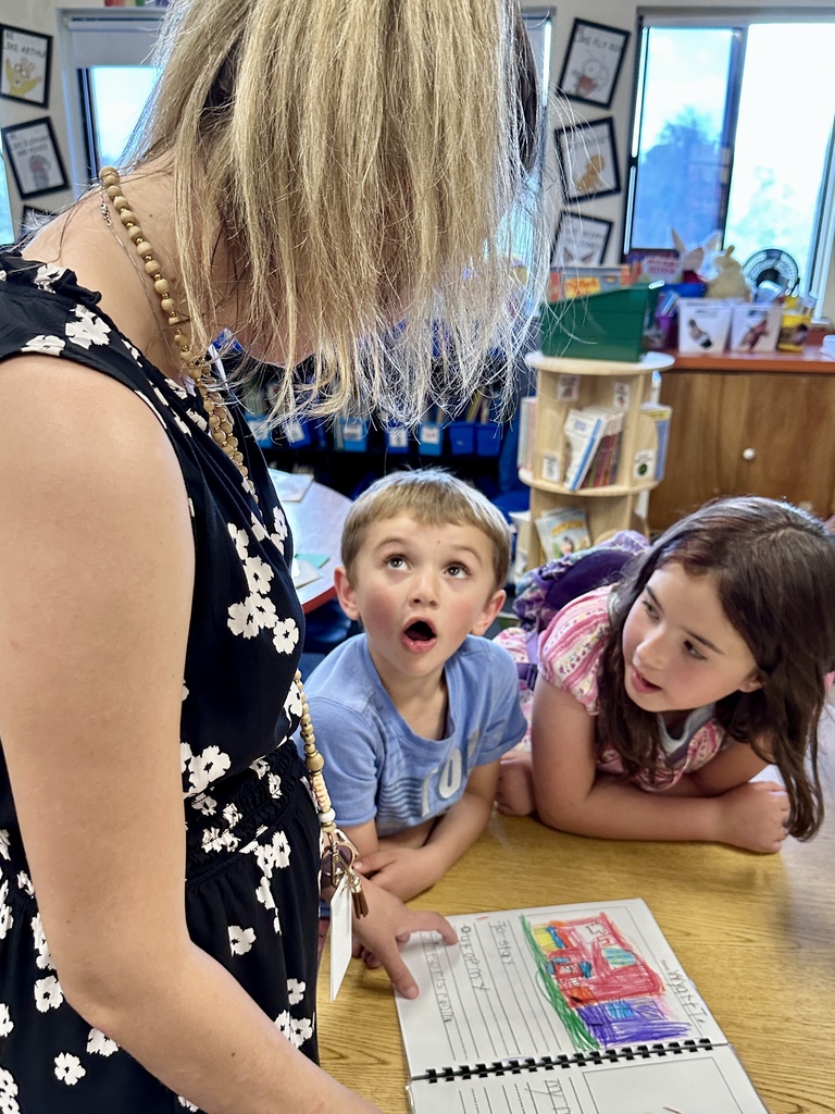Two students sit beside a teacher while proudly sharing a colorful illustrated project book at a classroom table.