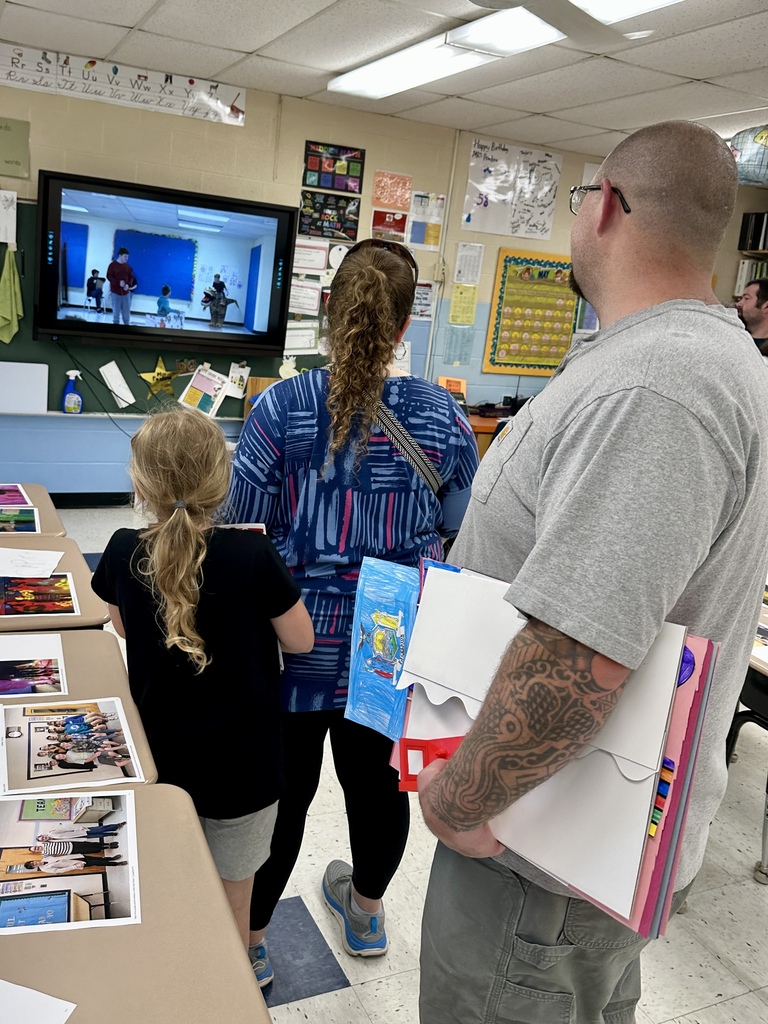 Family members stand in a classroom watching a presentation displayed on a classroom screen while holding student work and projects.