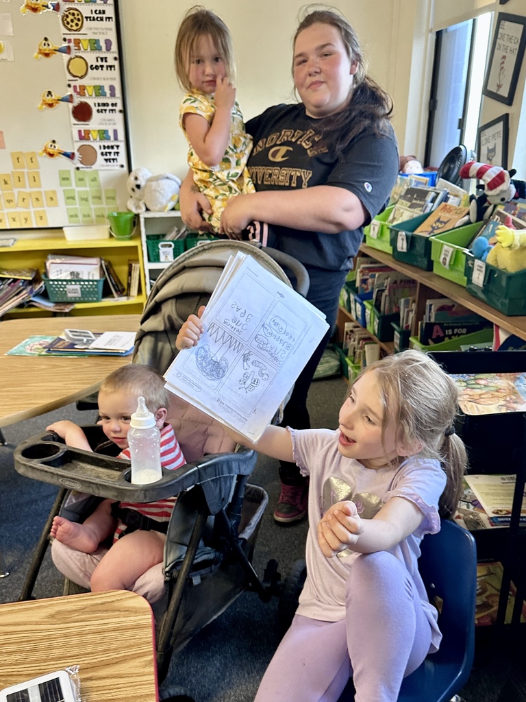 A student seated at a desk excitedly holds up work to show family members while a stroller and classroom materials are visible nearby.