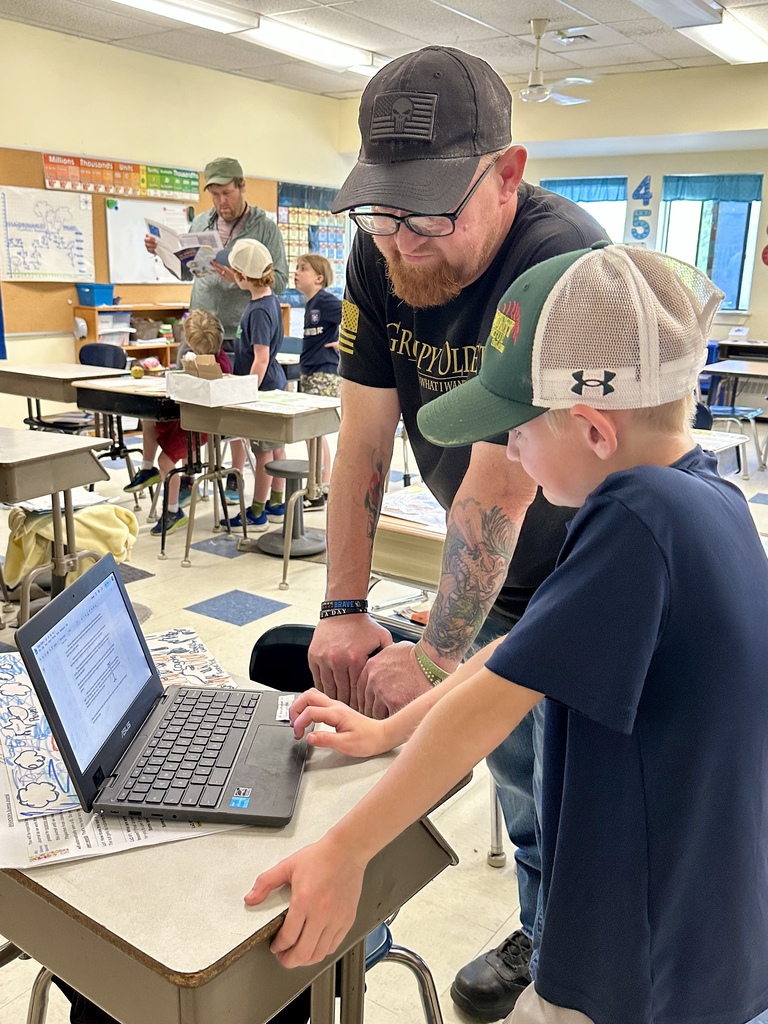 A parent leans over a desk beside a student while they work together on a laptop during classroom activities.
