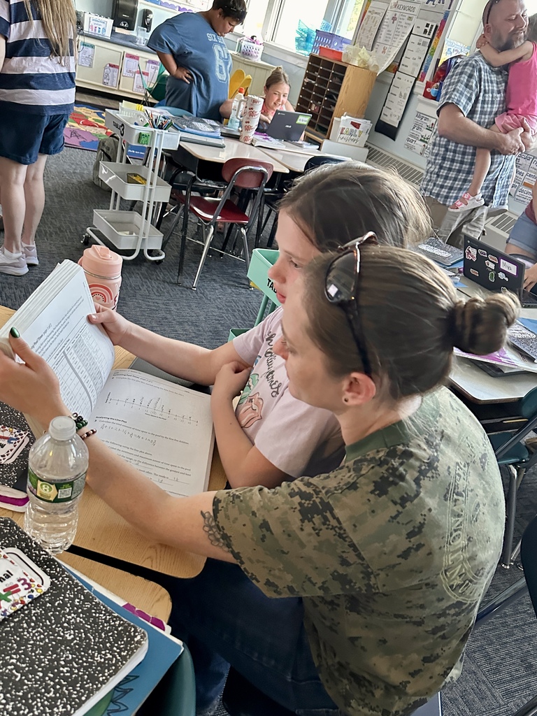 A family member sits beside a student helping review classwork and assignments together at a classroom desk.