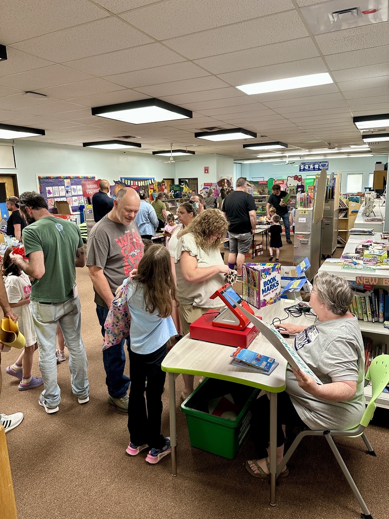 Families and students browse and shop at the Book Fair in the school library filled with books and displays.