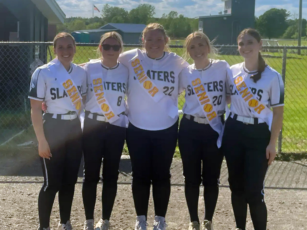 Five FHUHS softball seniors stand together on the field during Senior Night wearing white Slaters uniforms and gold “Senior” sashes. Pictured from left to right are Riley Babbie, Olivia Paige, Emma Surething, Victoria Genier, and Savannah Wilkinson smiling with their arms around one another in front of a chain-link fence and softball field on a sunny day.