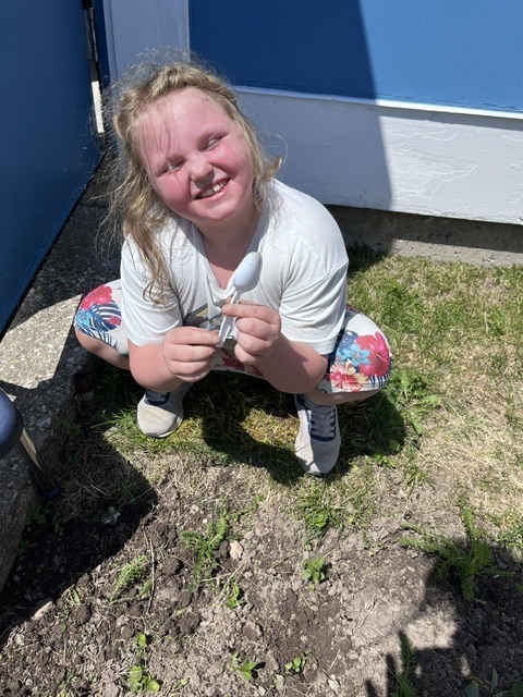 A young girl with light, wavy hair and a joyful expression squats down on a patch of grass next to a dirt garden bed. She is wearing a white t-shirt and floral-patterned shorts, and she holds a small white plastic spoon in both hands, smiling directly up at the camera.  image1.jpeg A group of young children are active outside on a sunny day next to a light-colored school building. Several children are crouched down, working in a long, narrow dirt garden bed that runs parallel to the building's wall. In the background, other children are running across a large, open green lawn under a blue sky with scattered clouds. 