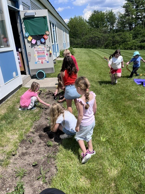  A group of young children are active outside on a sunny day next to a light-colored school building. Several children are crouched down, working in a long, narrow dirt garden bed that runs parallel to the building's wall. In the background, other children are running across a large, open green lawn under a blue sky with scattered clouds. 