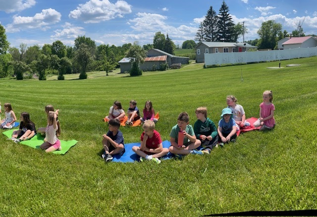 A wide, panoramic shot of a group of elementary school children sitting in a long line on colorful yoga mats laid out across a massive, rolling green field. The kids are facing forward on a bright, sunny day with a expansive blue sky filled with white, puffy clouds. In the far background, a house, barn, and tall evergreen trees line the edge of the property.